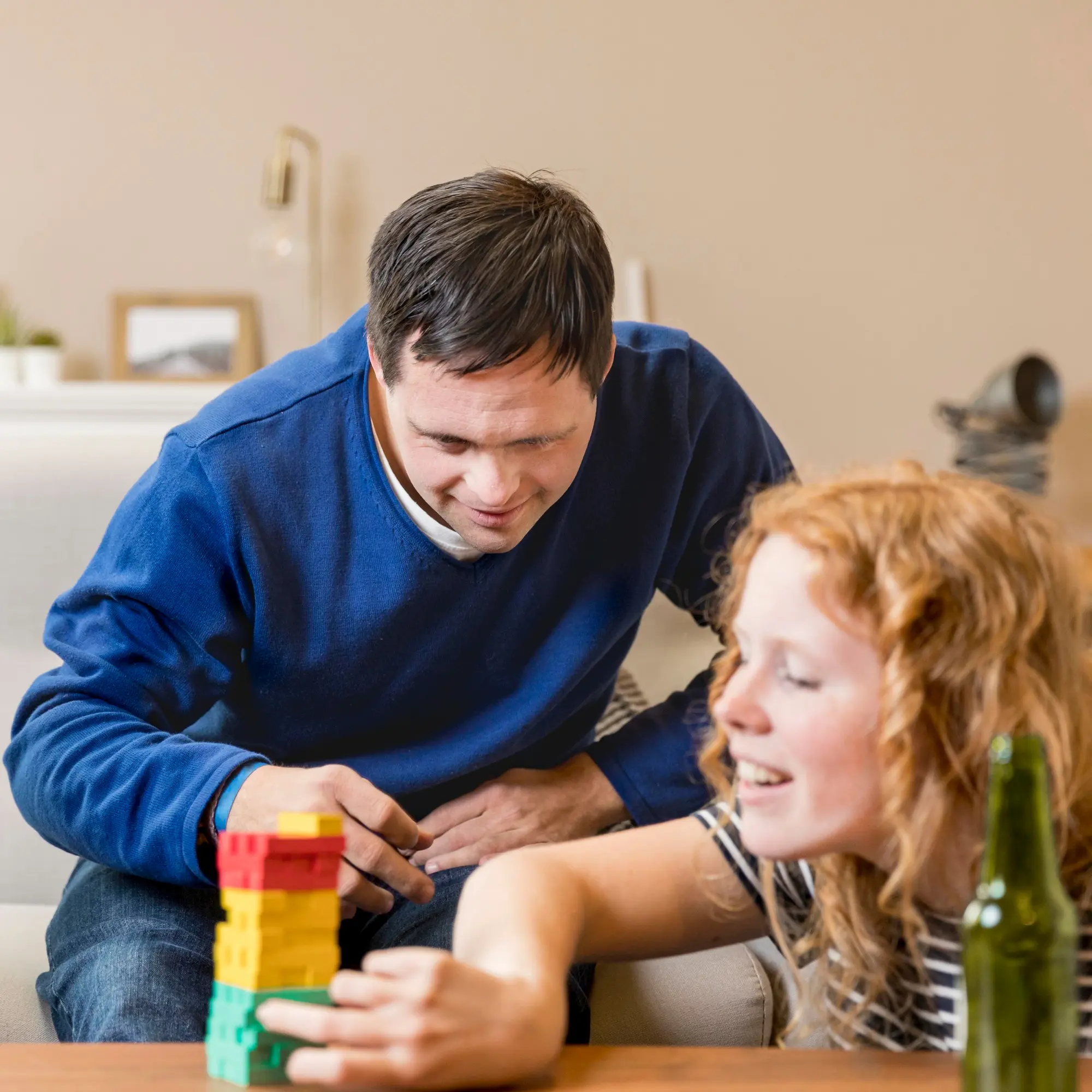 A man and a woman sit together on a couch, laughing while building a tower of colored blocks.