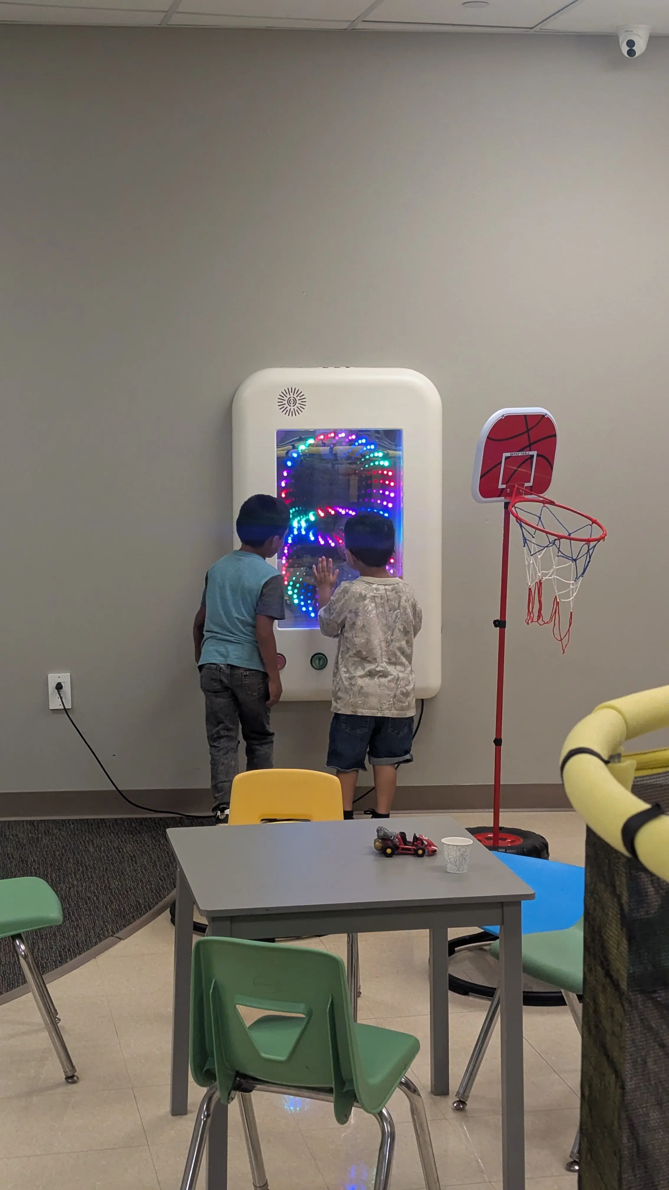 Two boys interact with a colorful lit sensory wall panel in a therapy play room.