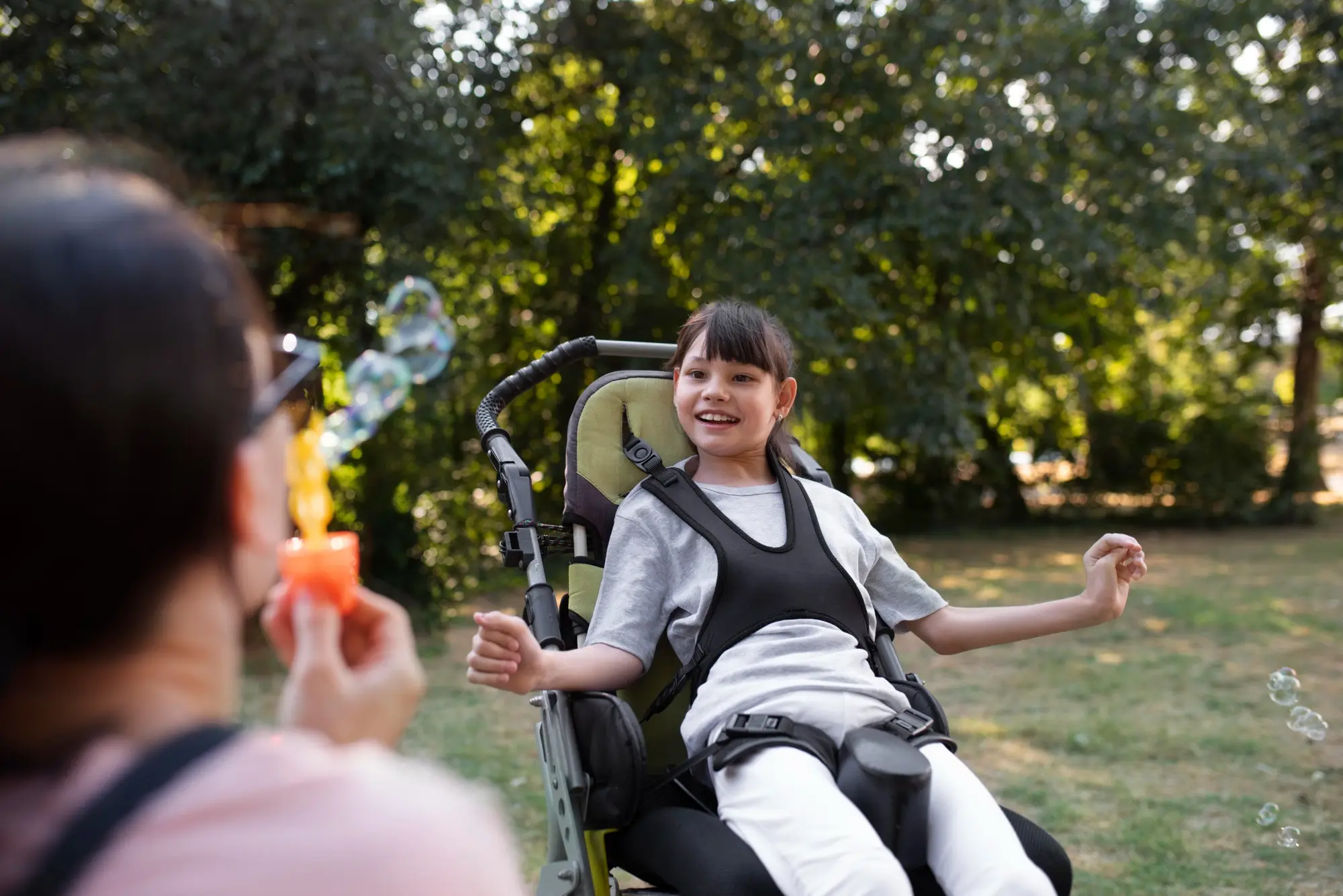 A caregiver blows bubbles for a smiling girl seated in an adaptive stroller in a sunlit park.