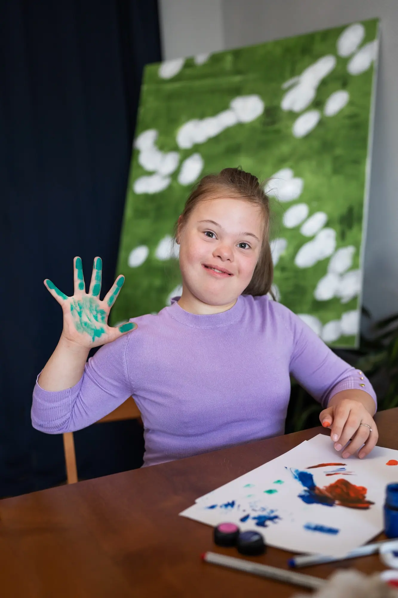 A girl in a purple sweater smiles and holds up a hand covered in green paint.