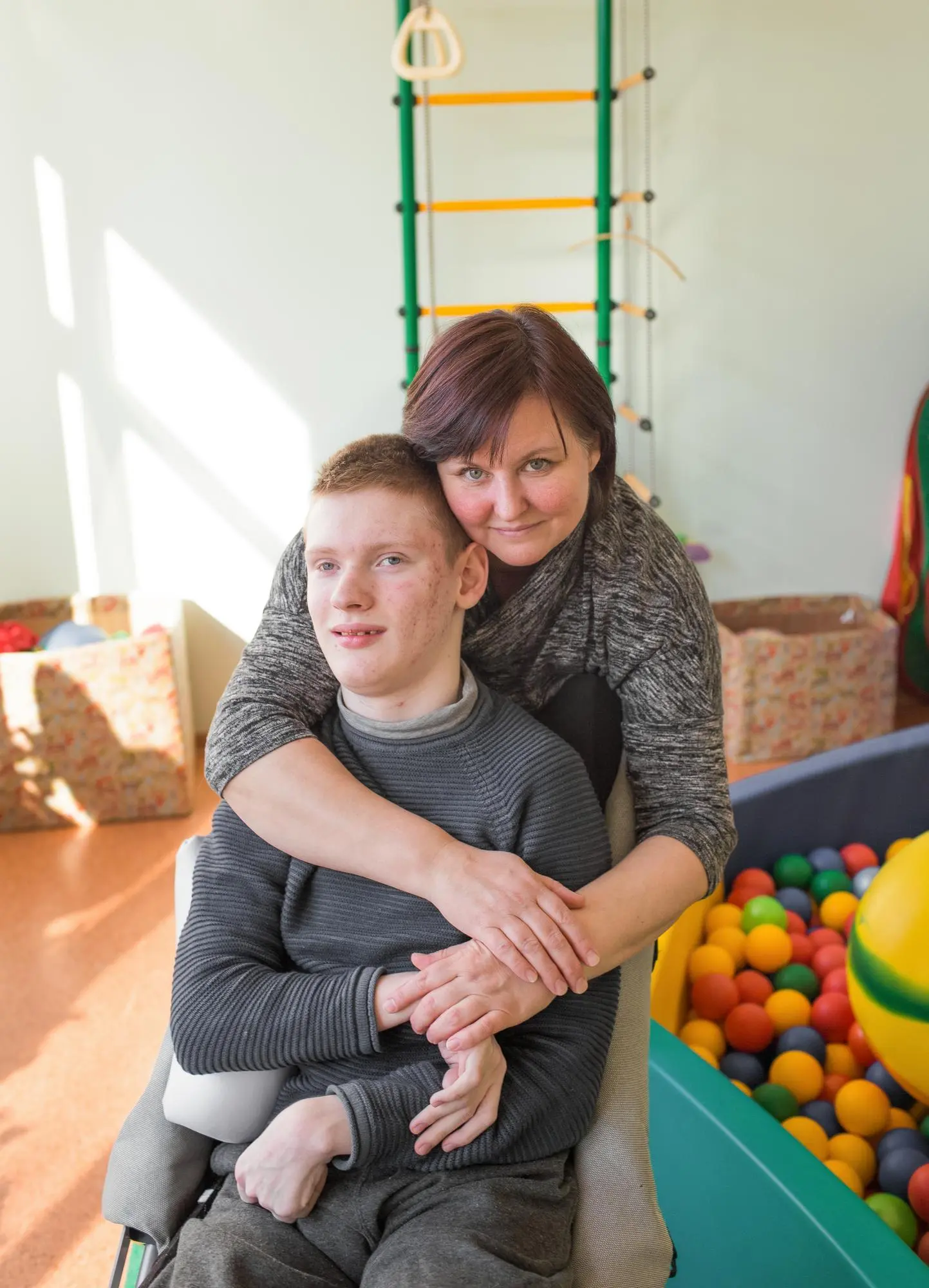 A caregiver leans down to hug a smiling young man seated in a sensory play room.