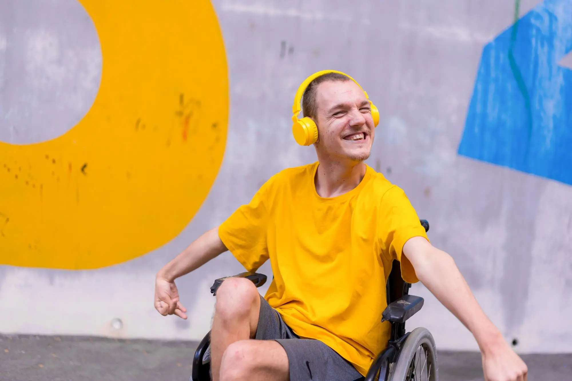 A young man in a yellow shirt and yellow headphones laughs joyfully in his wheelchair in front of a bright painted wall.